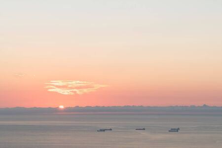 Minimalist shot of the sun peaking over the horizon over the water and boats. The sunrise clear sky has a blue pink gradient with a thin line of clouds over the sea in Rio de Janeiro, view from above.の写真素材