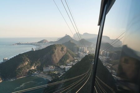 The aerial view from Sugarloaf Mountain over the buildings and city of Rio de Janeiro reflected in the window of the cable car. There are cables, Copacabana Beach and Praia Vermelha during the sunset.の写真素材