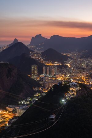 A pink sunset view at dusk over the night street lights of the city and buildings of Rio de Janeiro from Sugarloaf Mountain. A cable car travels from Morro da Urca. Botafogo, Pedra da Gavea are beyondの写真素材