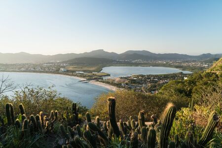 View over Itaipu Beach in Niteroi, Rio de Janeiro, from the Morro das Andorinhas hike. The early evening light hits the trees, cactuses, lake, white sand and mountains in the blue sky.の写真素材