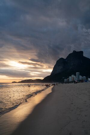 A cloudy sunset from Sao Conrado Beach in Rio de Janeiro, with the seaside granite mountain Pedra da Gavea. There are dark clouds with golden sunlight above the horizon shining in the sea water.の写真素材
