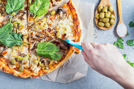 Delicious homemade pizza with tomato sauce, mushrooms, chicken, parmesan, peppers and olives on granite table. Top view, flat lay. Tasty Italian pizza. Man's hand cutting pizza with a roller knife.の写真素材