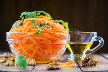 Bowl with fresh grated carrot. Organic salad with fresh carrot, walnuts, arugula and oil in glass bowl on wooden background. Healthy food, detox salad. Diet concept.の写真素材