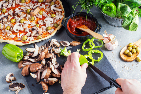 Pizza preparation, raw dough surrounded by ingredients. The man cuts mushrooms and peppers for pizza. Male hands cut the pepper on a slate board.の写真素材