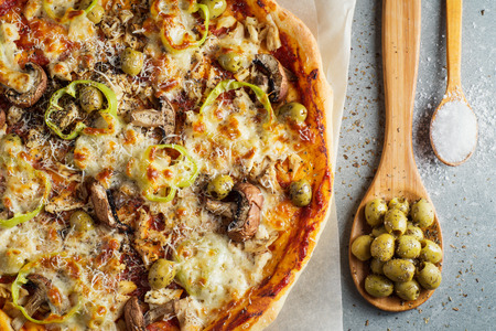 Delicious homemade pizza with tomato sauce, mushrooms, chicken, parmesan, peppers and olives on the granite table and wooden spoons with olives and salt. Top view, flat lay. Tasty Italian pizza.の写真素材