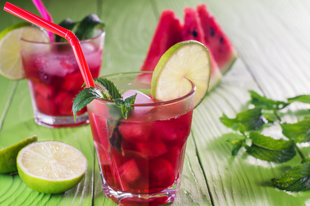 Watermelon cocktail in two glasses with slices of watermelon, lime, mint and ice, melon mojito. Homemade lemonade with watermelon and red striped straw on green wooden table. Summer drink.の写真素材