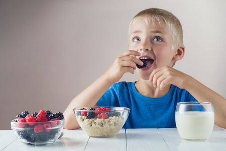 Happy, smiling child eating oatmeal with berries. The concept of a healthy breakfast for children, porridge with raspberries and blackberries, a glass of yogurt on white wooden table.の写真素材