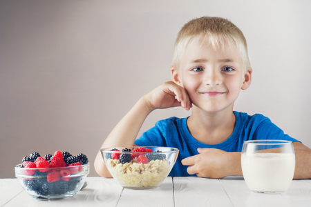 Happy, smiling child eating oatmeal with berries. The concept of a healthy breakfast for children, porridge with raspberries and blackberries, a glass of yogurt on white wooden table.の写真素材