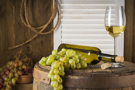Still life of white wine with wooden keg with copy space. Wine bottle, glass of white wine and bunch of grapes on a old wooden barrel.  Wine tasting and production concept.の写真素材