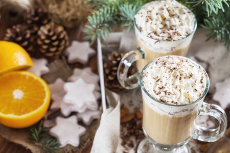 Christmas background with two latte, cookies star, fir branches, cones, orange and spices, close up. Couple coffee with milk and homemade tasty cookies in tray on wooden background. Top view.の写真素材