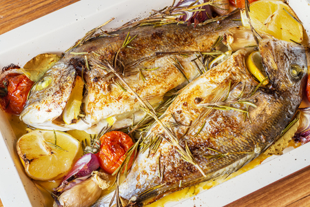 Two delicious whole baked fish. Baked dorado with lemon, onions, rosemary, cherry tomatoes, and spices on a wooden background. Delicious roasted dorado on oven tray. Diet and healthy food. Top view.の写真素材