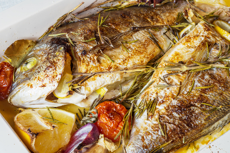 Two delicious whole baked fish. Baked dorado with lemon, onions, rosemary, cherry tomatoes, and spices on a wooden background. Delicious roasted dorado on oven tray. Diet and healthy food. Top view.の写真素材
