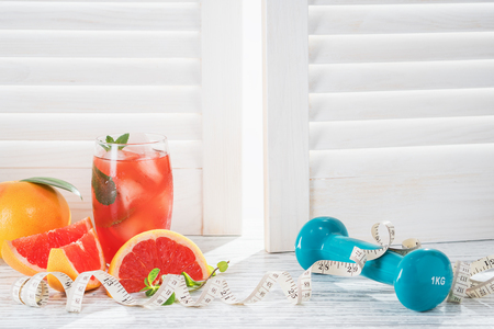 Fresh grapefruits and grapefruit juice, dumbbell and measuring tape, on rustic white wooden table opposite the blindsの写真素材