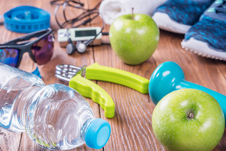 Set for sports activities on brown wooden background. Sport equipment, sport shoes, measuring tape, dumbbell, hand expander, towel, apples and bottle of waterの写真素材