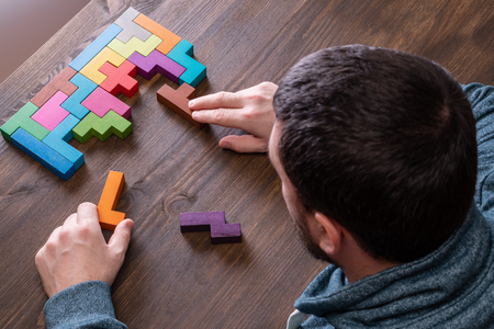 A man connects different geometric shapes, the concept of concentration, logical thinking. Top view on man's hand playing with colorful wooden blocks on the brown wooden table background.の写真素材