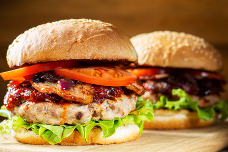 Two fresh tasty burger. Homemade hamburger with fresh vegetables, close-up. Sesame buns with succulent beef patty, grilled onions, bacon, tomato and fresh salad served on rustic wooden board.の写真素材
