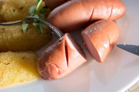 Pork sausages and breaded potatoes fried in a dish.  Fork and knife placed next to dish on wooden tableの写真素材