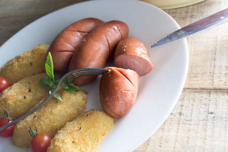 Pork sausages and breaded potatoes fried in a dish and decorated with cherry tomato.  Fork and knife placed next to dish on wooden tableの写真素材