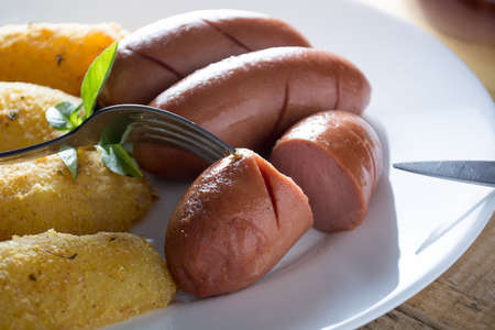 Pork sausages and breaded potatoes fried in a dish.  Fork and knife placed next to dish on wooden tableの写真素材