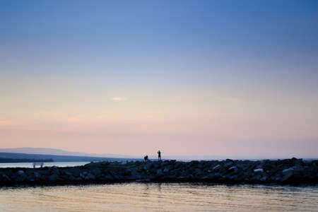 Sea barrier made of stones in the sunset with two man silhouettes on itの写真素材