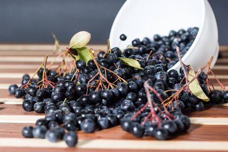 White bowl full with fresh and tasty aronia spilled on wooden table
