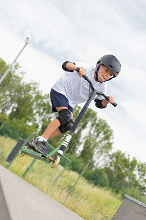 The boy jumps on a scooter. A cute little boy rides a scooter in a skatepark. A young novice athlete spends free time in extreme sports.の写真素材