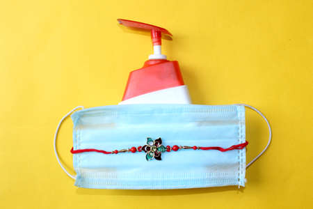 Raksha Bandhan Festival concept in Corona Virus or Covid-19, traditional Rakhi or Wrist Band bracelet with sanitizer and protective mask for precaution over on yellow background.の写真素材