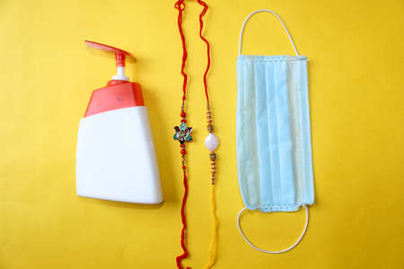 Raksha Bandhan Festival concept in Corona Virus or Covid-19, traditional Rakhi or Wrist Band bracelet with sanitizer and protective mask for precaution over on yellow background.の写真素材