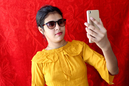 Beauty fashion portrait. Smiling young woman in sunglasses mobile selfie pose and Wear yellow top, fashion photo on red background, selective focus with blur.の写真素材