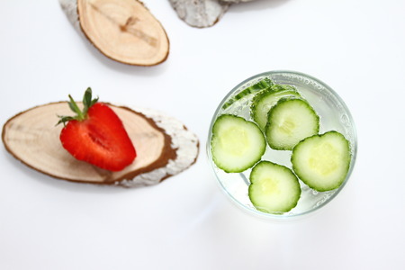 Detox flavored water with cucumber on white background with red strawberry and wood decoration. Healthy food concept.  Refreshing summer homemade cocktail. Copy space. No sharpen. Anti stress drink.の写真素材