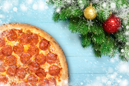 Christmas Pizza pepperoni with snowflakes and fir branches, new year toy ball or bauble on blue wooden background. Winter holiday decoration top view.の写真素材