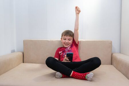 The boy in the red shirt sitting at home on the couch playing on your phoneの写真素材