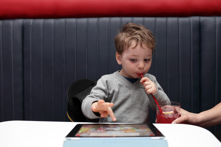 Child drinking juice and playing on tablet PC in the cafeの写真素材