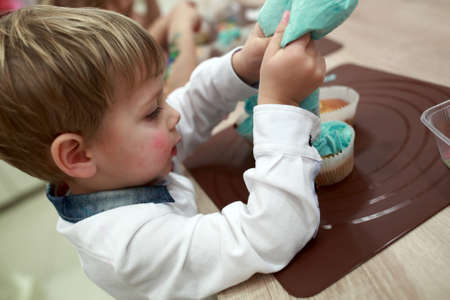 Child decorates cupcakes with cream in the kitchenの写真素材