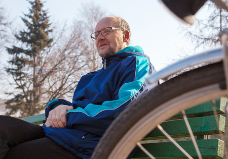 portrait of eldely man wearing glasses, black trousers and a blue jacket sitting on a bench near his bicycle in a city parkの写真素材