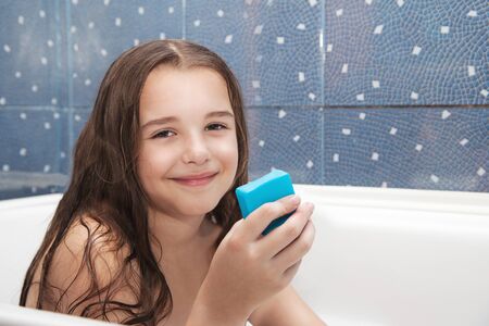 little smiling girl with long brown hair holding soap sitting in the bathの写真素材