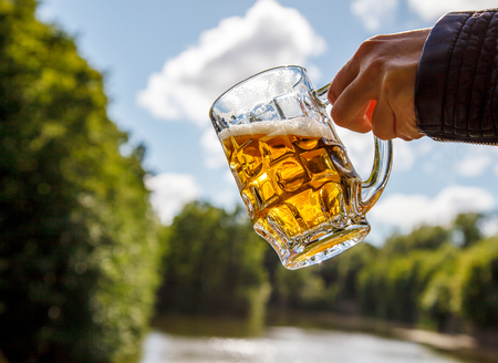 female hand holding a mug of beer near the lake on sunny summer dayの写真素材