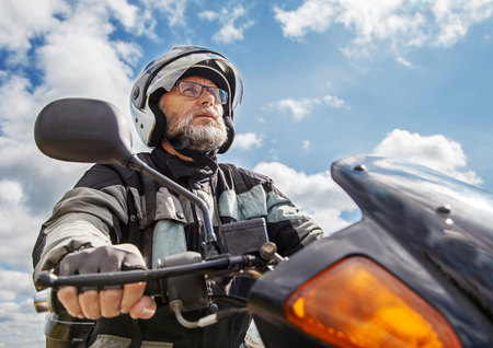 elderly motorcyclist wearing a jacket and glasses with a helmet sitting on his motorcycle on the road closeupの写真素材