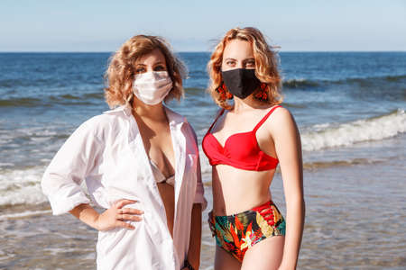 two young blonde girls in a swimsuits and protective masks standing on the seashore on sunny summer day closeupの写真素材