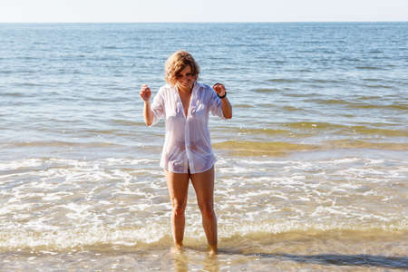 young blonde woman in a wet white shirt standing in the water near the seashore on sunny summer day