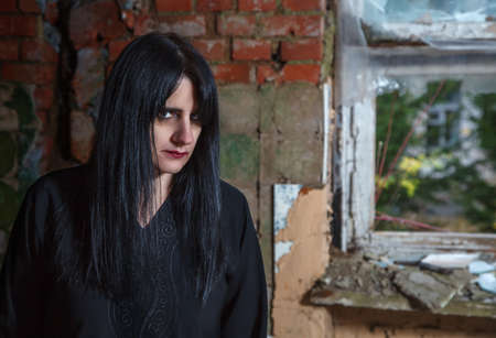 portrait of young beautiful goth woman in black dress standing near broken window in an abandoned...