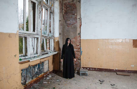 young beautiful goth woman in black dress standing near broken window in an abandoned house