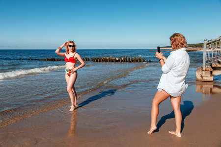 two girls in swimsuits take pictures of each other with smartphones on the seashore on sunny summer dayの写真素材