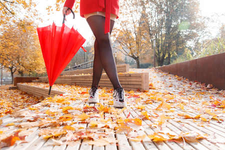 woman in red coat and umbrella standing on alley of city park on sunny autumn day. legs closeupの写真素材