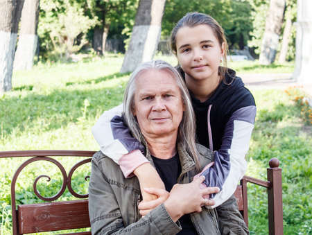 granddaughter hugs her grandfather on a bench in a city park on sunny autumn dayの写真素材