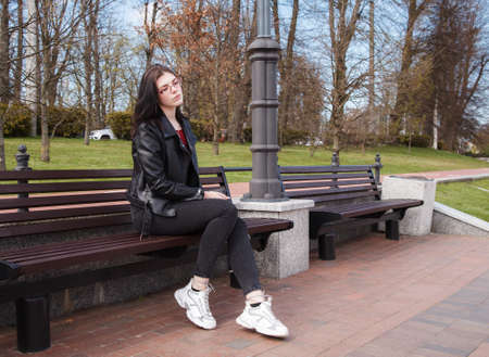 young girl in black jacket and jeans sits on a bench in a city park on sunny spring dayの写真素材