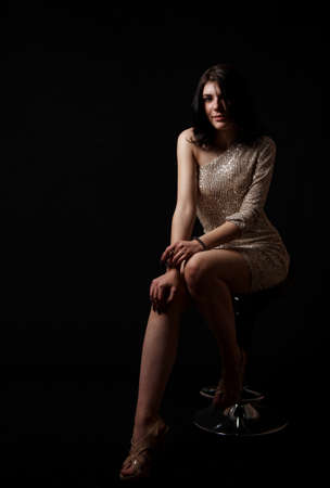 young beautiful brunette girl in bright dress posing sitting on chair in the studio on black backgroundの写真素材