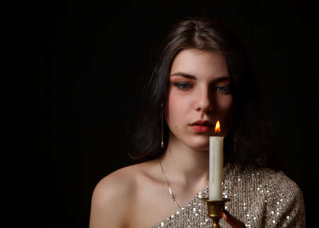 young beautiful brunette girl in bright dress holding burning candle in studio on black backgroundの写真素材
