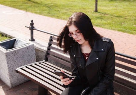 young girl in black jacket and jeans looking at smartphone display while sitting on bench in city park on sunny spring day. outdoor closeupの写真素材