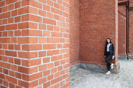 young beautiful brunette girl in black jacket and jeans stands near wall of gothic church on summer dayの写真素材